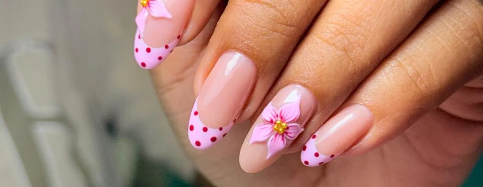 A client's hand with almond nails in a soft pink, tipped with pink and red polka dots and a sculpted flower on the ring finger